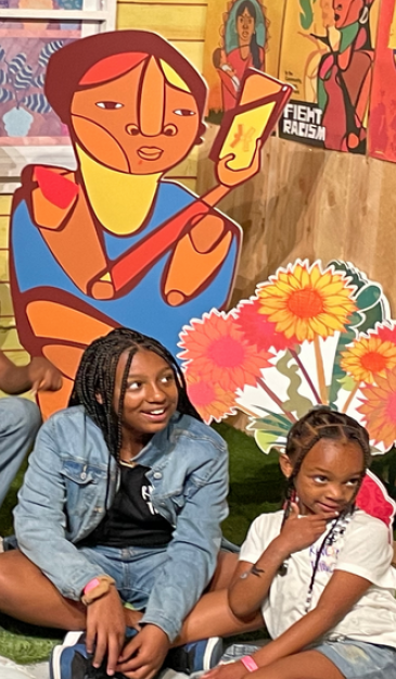 Two female black youth sitting and smiling in front of a colorful art installation